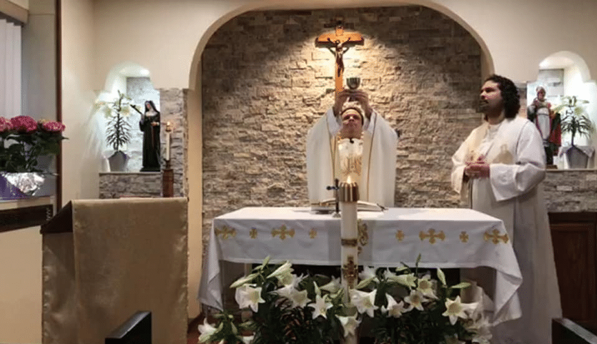A priest stands behind a table in a church, offering mass. Another priest stands beside him.