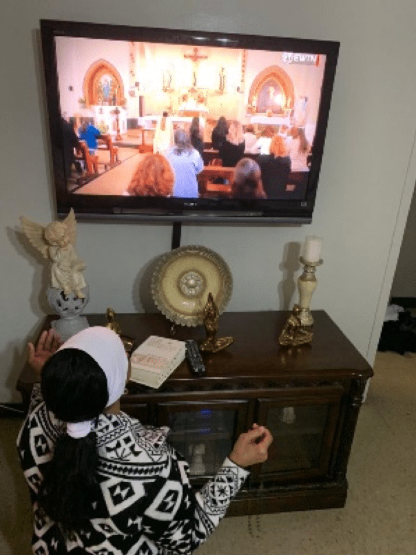 A woman -holding a  Rosary and kneeling - watches Mass on her TV.