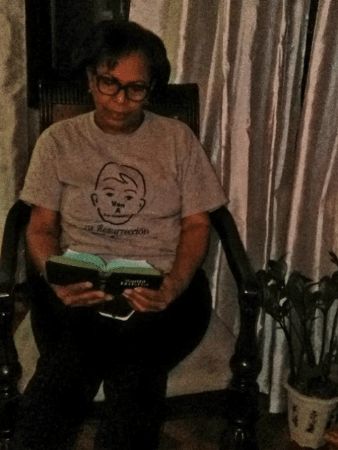 A woman (Pura Rodriguez) sits in a chair and reads her Bible at home.