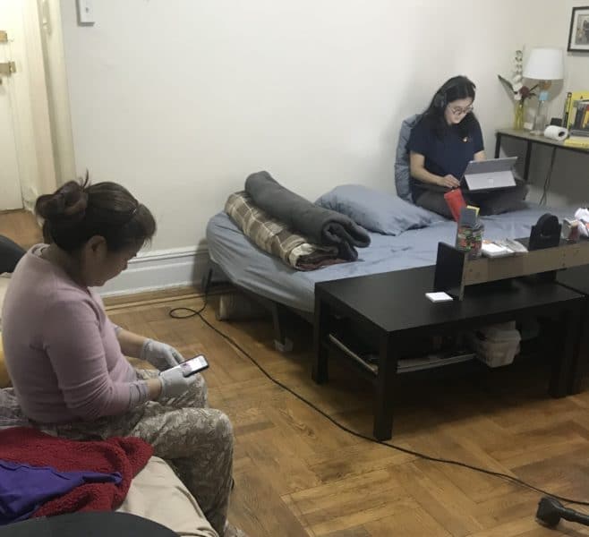 A picture of the author working on her computer on her bed in the  corner of the family living room. Another family member wears sanitary gloves and sits on the couch looking at her phone.
