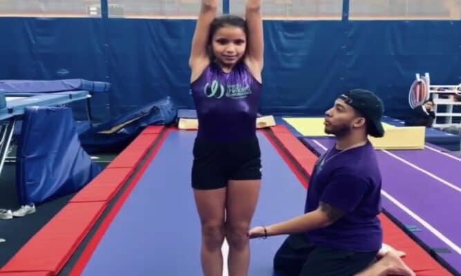 A gymnastics coach kneels and helps a young, female gymnastics student