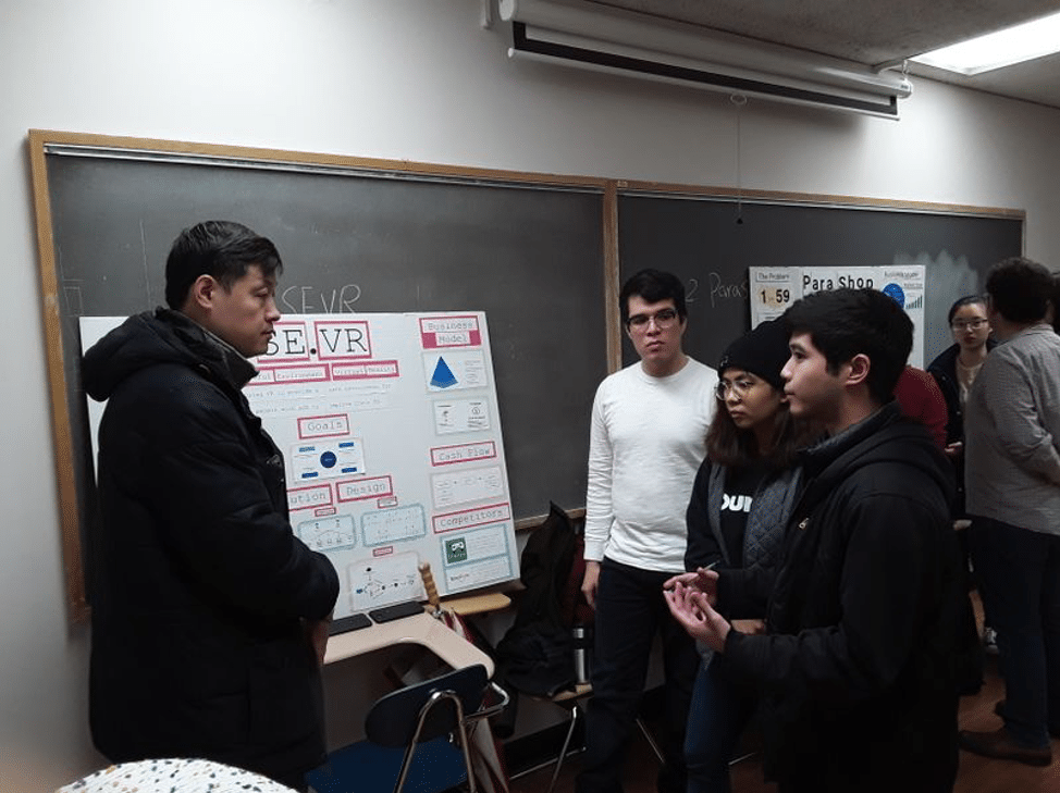 Four people stand in a classroom looking at a poster board entitled "SE.VR" (an early title for Undaunted). The four people are advisor Hao Tang, and teammates: Carlos Saravia, Abigail Banting, Raymond Rodriguez.