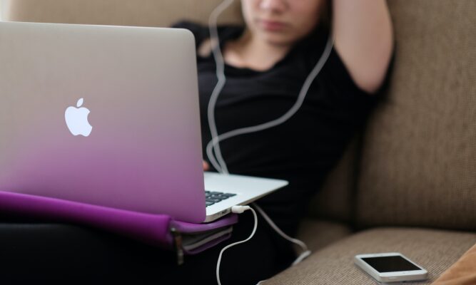 Female student on a couch with open laptop on her lap and earbuds in ears.