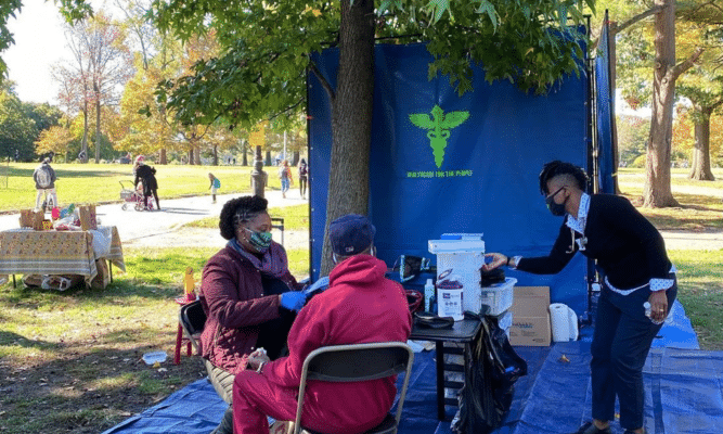 Two health care providers gather around a patient who is sitting in a chair in a make-shift medical station in Prospect Park. Blue tarp is on the ground. Blue sign with a medical sign in the background.