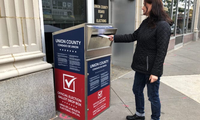 A young woman wearing a black coat and black pants drops off her mail-in ballot.