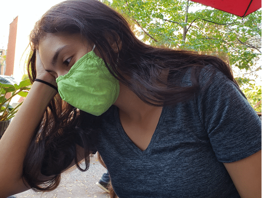 A woman wearing a green mask rests her head on her hand and looks pensive.