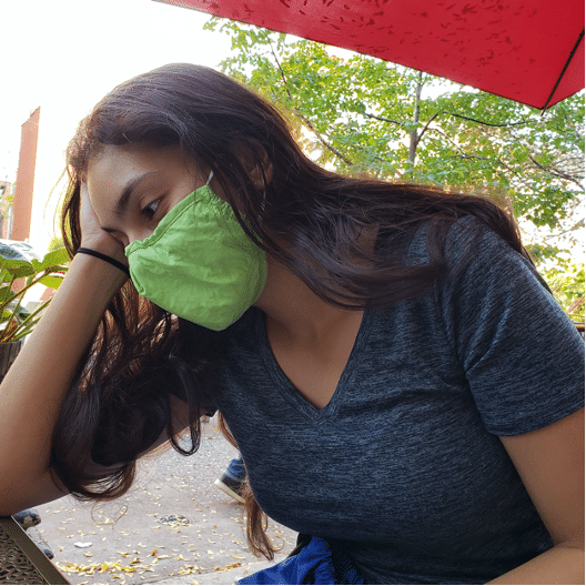 A woman wearing a green mask rests her head on her hand and looks pensive.