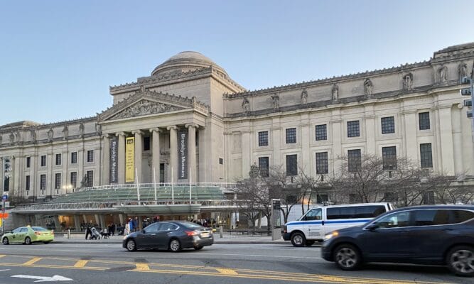 Photo of the front of the Brooklyn Museum with some cars passing in front.