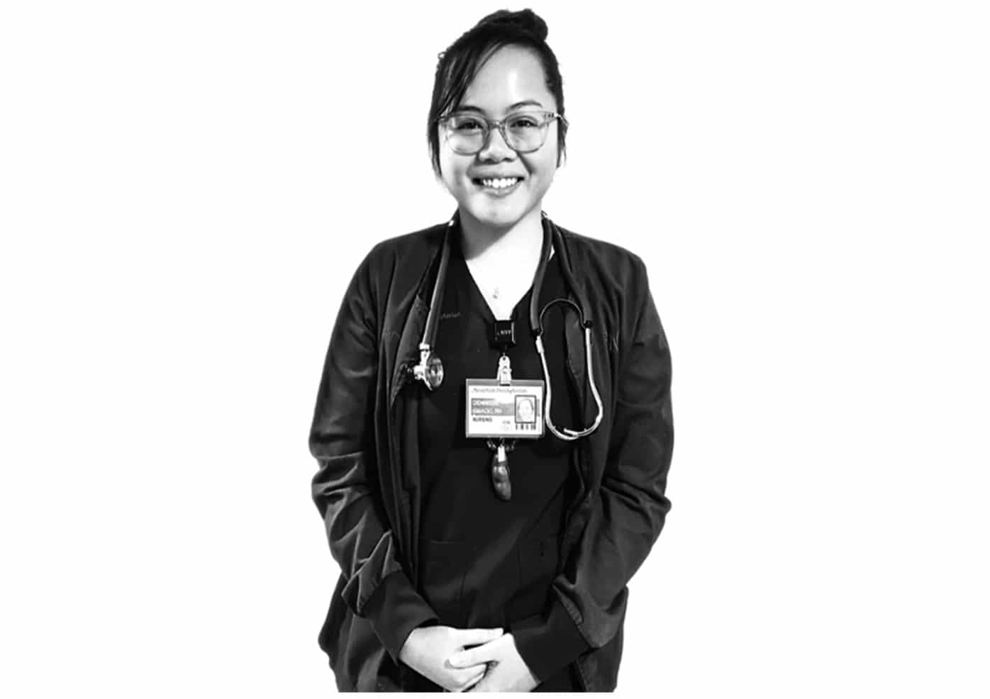 A black and white photo of Dennisse Carla Gejon in her nurse's scrubs and a stethoscope and I.D. around her neck - smiling directly at the camera. 