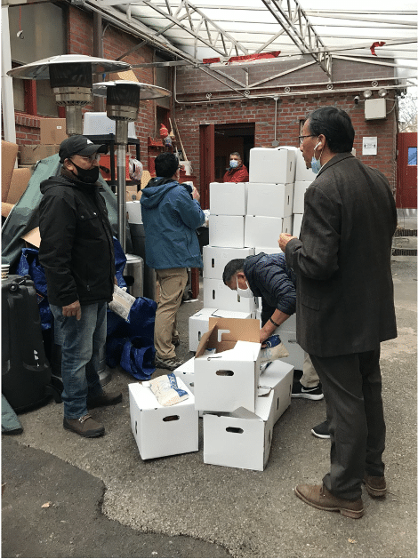 Urgen Sherpa and other members of the association are shown outside in winter coats and COVID masks as they discuss the pictured dozens of stacked white boxes that are left over from a food drive.