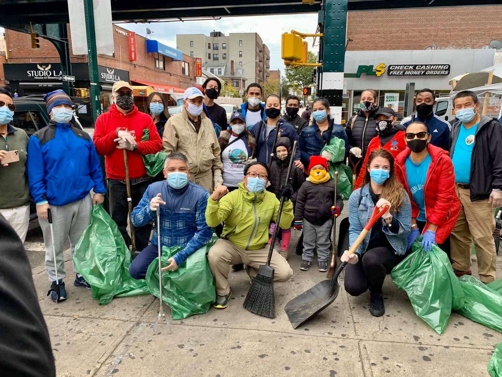 Photo shows about 20-22 people posing with shovels, rakes, and brooms and holding green trash bags. They are all wearing COVID masks and have coats on. There are men, women and young children included. 