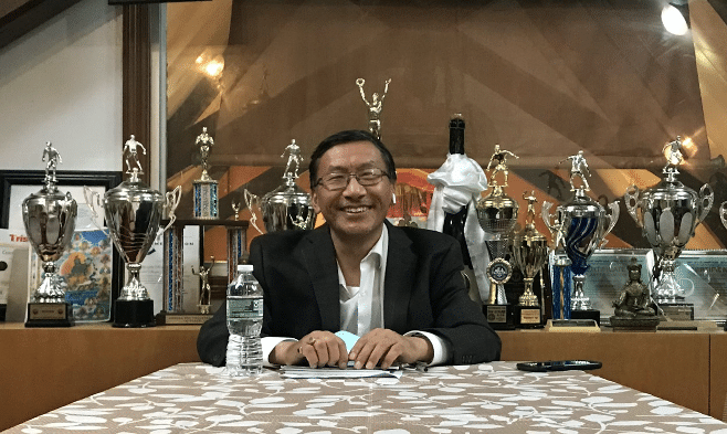 Urgen Sherpa is seated at a table and behind him are about 15 large trophies. Mr. Sherpa is smiling and seems very happy. He wears a white dress hirt and a dark colored sports jacket. His hands are on some papers. He has a disposable water bottle beside him and his cell phone is lying on the table to his left (our right). 