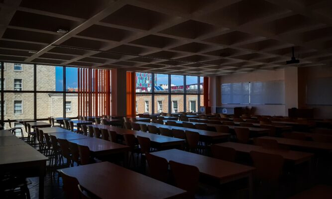 Picture shows a dimly-lit (lights off) classroom filled with at least 24 4-6-foot tables with chairs. The room is completely empty. There are white boards at the front of the room and large windows overlooking part of a school building. It's a sunny day outside with blue skies.