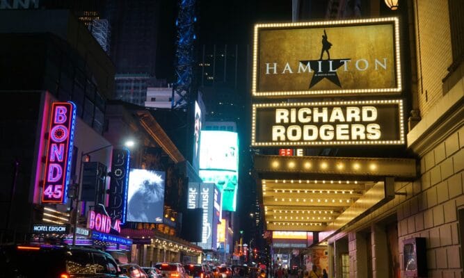 A street in the theater district in NYC before the lockdown showing a lit sign for the Richard Rodgers Theater where Hamilton was showing. The photo shows other lit theaters and buildings down the street. It's a nighttime view of a busy NYC street.