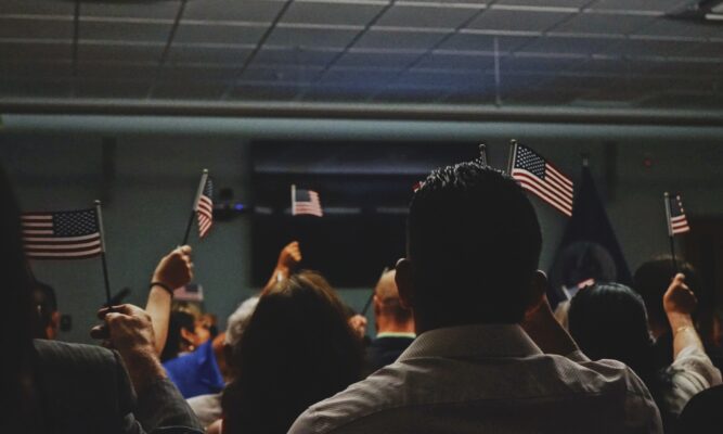 We see the backs of a room full of people who are holding miniature American flags.