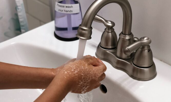 white bathroom sink, silver faucet, clear hand sanitizer jar, two hands and forearms seen. Hands are under running water as if handwashing.