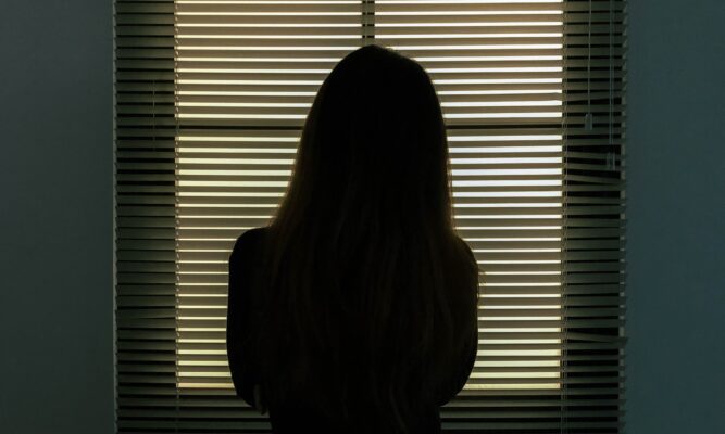 silhouette view of the back of a woman with long hair looking out a window with blinds over the window.
