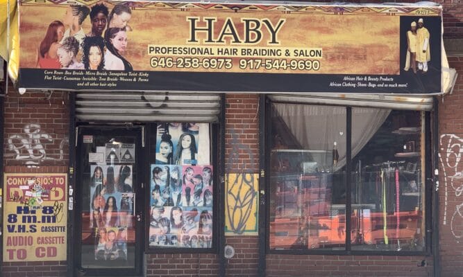Picture of a storefront of the braiding shop. The sign says: Haby Professional Hair Braiding & Salon. The sign has yellow, orange, red tones. It includes pictures of women with different braids. The store has a brick front. one level. There are more photos of women with braids on the door.