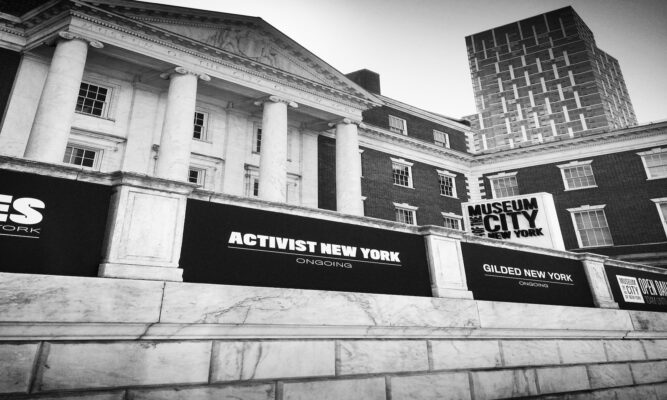 a black and white photo of the front of the Museum of the City of New York - the front center of the building is white and has columns. We can see a side wing that is brick. There are black banners along some sort of concrete "fence" that say things like: "Activists New York Ongoing" and "Gilded New York."