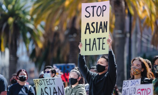 Picture shows about 11 people - of a bigger crowd - mainly Asian - protesting and holding signs. The predominant sign, held high by an Asian man, says "Stop Asian Hate." A couple of other signs that we don't see in their entirety, say, "Protect Asian ..." (the end of the slogan is unseen) and "Stop" and "Bad Days Don't Lead to Murder."