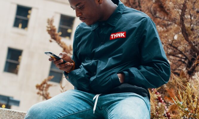 A male presenting dark skinned individual, approx. 20-30 years old, wearing blue jeans and a dark light-weight jacket that says “Think” on the upper-left hand side, sitting outside on a stone wall, stares at his phone, which he holds in his right hand. His left hand is in the left coat pocket. We ca see windows from a large white building behind him. It could be a college or an office building. There’s a tree behind them with not-vibrant fall leaves.