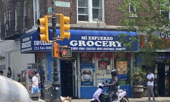 a street view photo of a Dominican bodega called Mi Esfuerzo located in upper Manhattan on Dyckman Street. We see a couple of people outside on the sidewalk and a person sitting on a motorbike in front of the store.