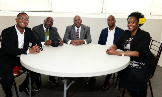 5 Black people - 4 men and 1 women, sit around a round table - looking at and smiling at the camera.
