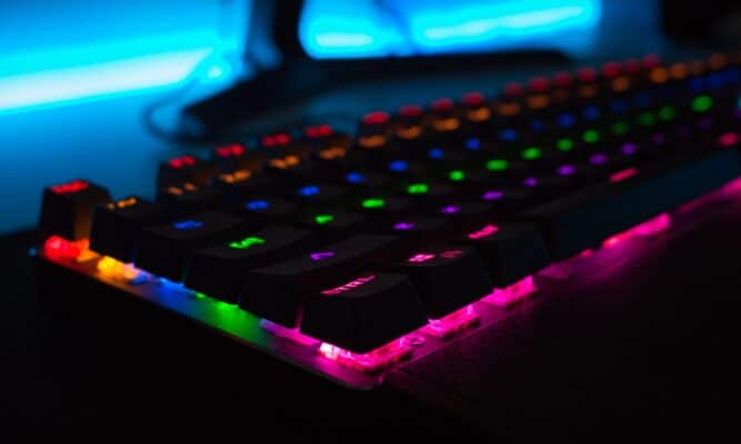 a close up picture of a computer keyboard - dark and moody - looking from the bottom left corner. Keyboard is dark and the sides and letters are illuminated in red, blue, green and pink. We don't see the entire keyboard.