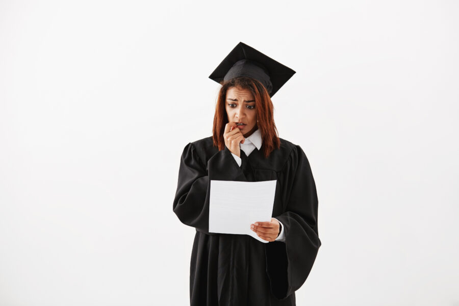 Sad displeased african female graduate in black mantle holding test over white background. Copy space.