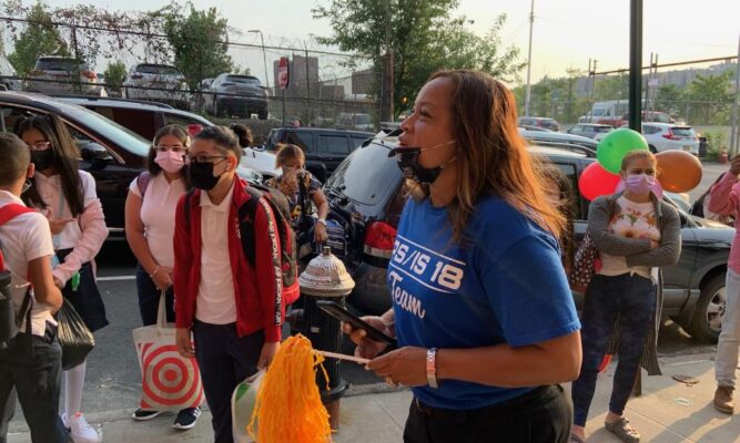 A woman with brown skin, the principal of the school, is holding an orange pom-pom in one hand and her phone in the other. She's wearing a blue, short-sleeved t-shirt that says PS/IS 18 Team, is standing outside on a sidewalk talking to students of various ages and ethnicities. The day is clear. There are both parked and moving cars on the street behind the students.