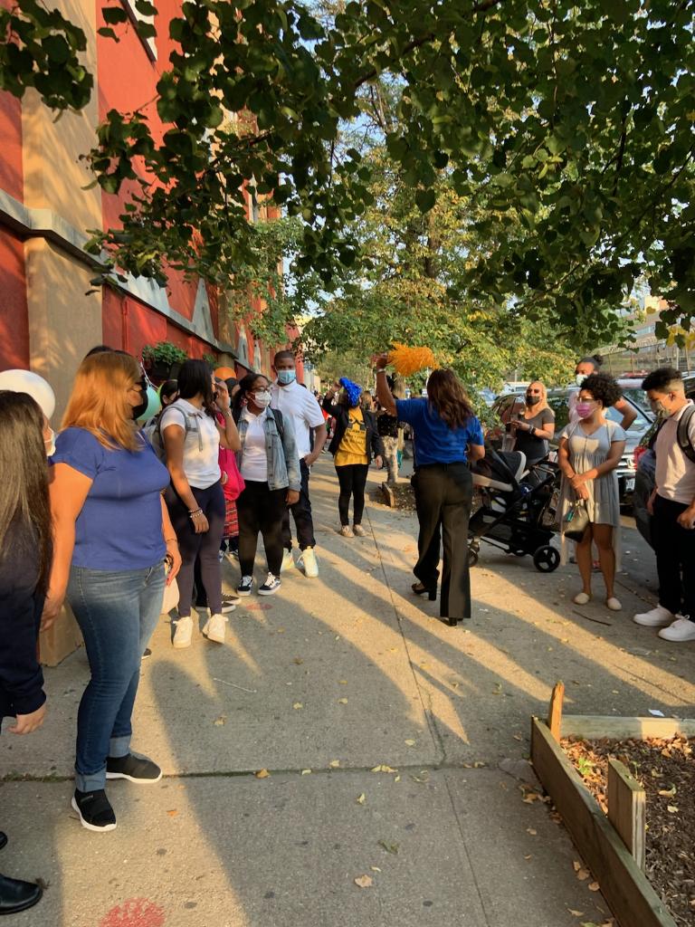 Students line the sidewalk and principal Connie Mejia walks between them, holding an orange pom pom and welcomes them back to school.