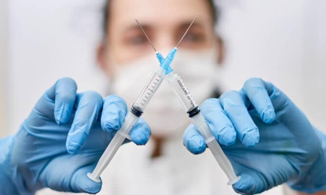 A masked medical professional with blue medical gloves holds a syringe in each hand and they're crossed making an "x" - indicating no vaccine.