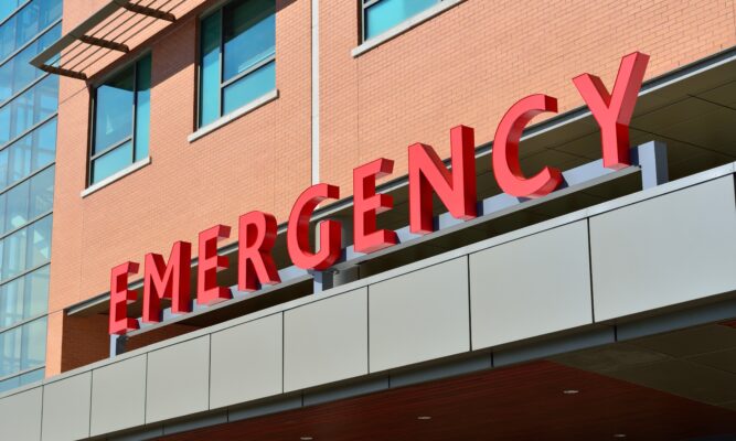 A photo of a red EMERGENCY sign depicting the ER entrance of an orange brick hospital