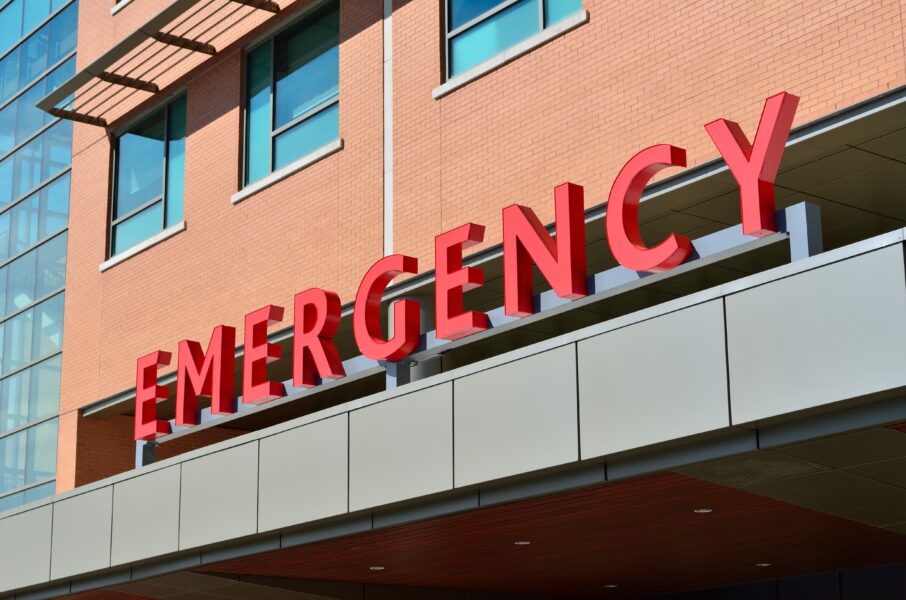 A photo of a red EMERGENCY sign depicting the ER entrance of an orange brick hospital