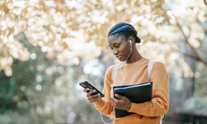 A Black female-presenting college student is outside. A tree is behind her. She's looking at her phone and she has a slight smile. e