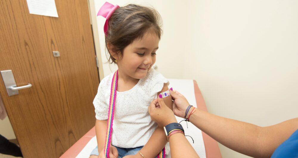 Little girl sits on doctor's office exam bed while hands of a medical professional puts a bandaid on her arm.
