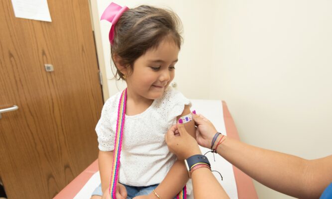 Little girl sits on doctor's office exam bed while hands of a medical professional puts a bandaid on her arm.