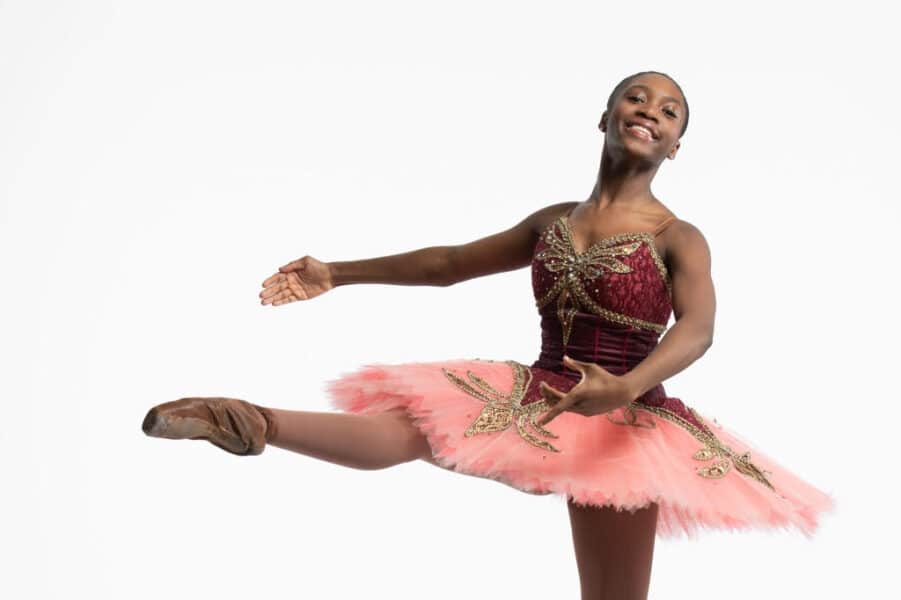 a young woman in a burgundy and pink leotard and tutu smiles and holds a dance pose with her right leg extended in front of her.