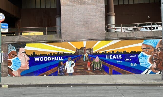 A mural on a brick wall of the outside of Woodhull Hospital that says Woodhull heals and has images of health care workers and patients. The word “Heals” is written in several languages. 