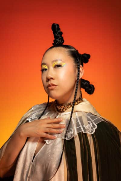 A woman with three buns in her hair poses with her right hand on her chest.