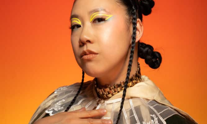 A woman with three buns in her hair poses with her right hand on her chest.