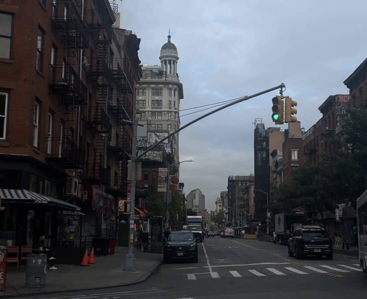 The corner of a NYC street in the lower east side with a somewhat busy street and an empty sidewalk.