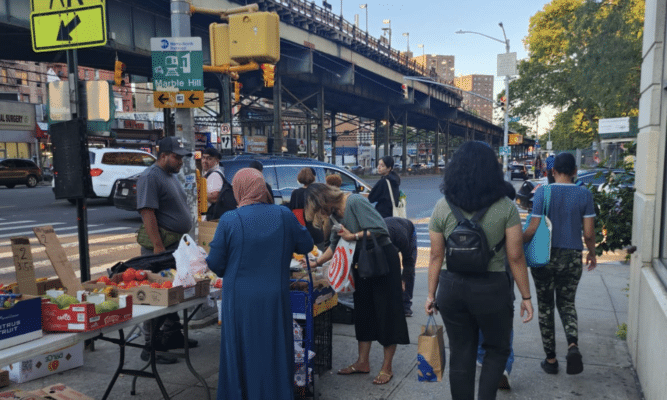 A group of pedestrians on a sidewalk, photographed from behind. A few women stop at a produce stand in the left of the frame.