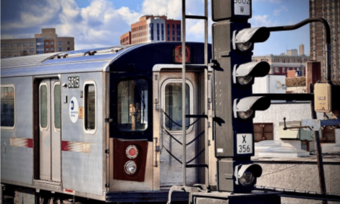 An above-ground train in New York City. The train car comes from the left side of the frame and the blue sky peaks over the top of it.