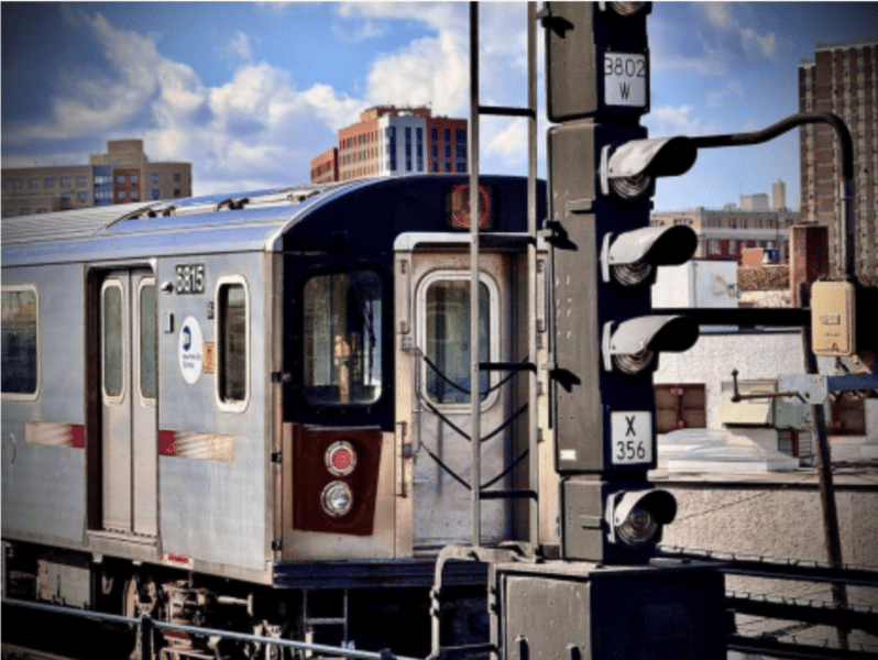 An above-ground train in New York City. The train car comes from the left side of the frame and the blue sky peaks over the top of it.