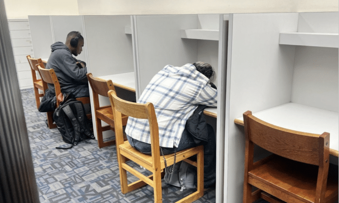 The photo depicts two students at gray desks in a library. One student wears headphones and appears to be working. The other has their head down on a desk and appears to be napping.