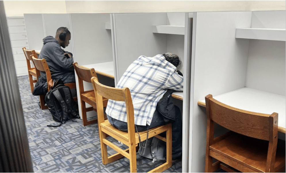 The photo depicts two students at gray desks in a library. One student wears headphones and appears to be working. The other has their head down on a desk and appears to be napping.