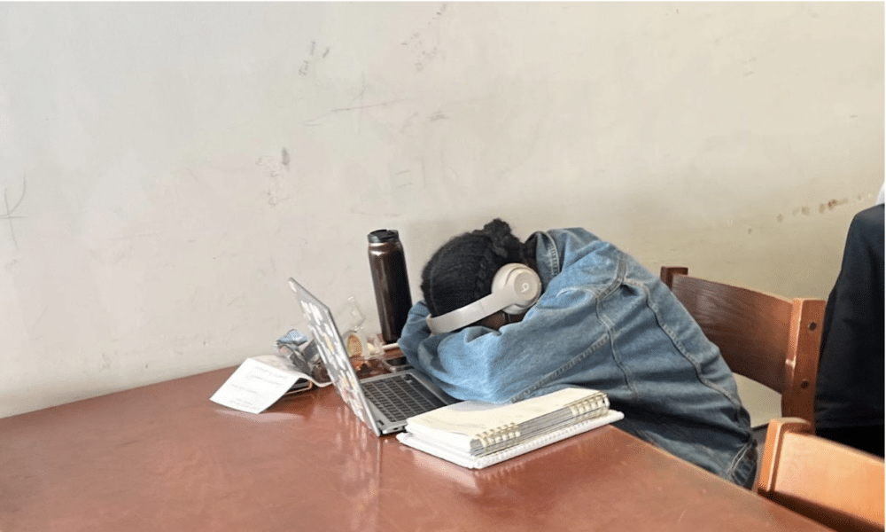 A student wearing headphones rests her head on a desk in front of an open laptop and notebook. 