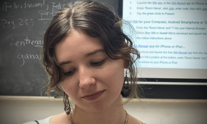 A brunette woman with earrings and a necklace looks down into the bottom left corner of the frame. There is a blackboard in the background.