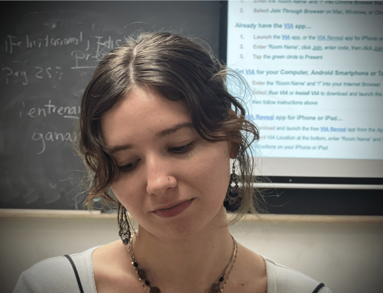 A brunette woman with earrings and a necklace looks down into the bottom left corner of the frame. There is a blackboard in the background.